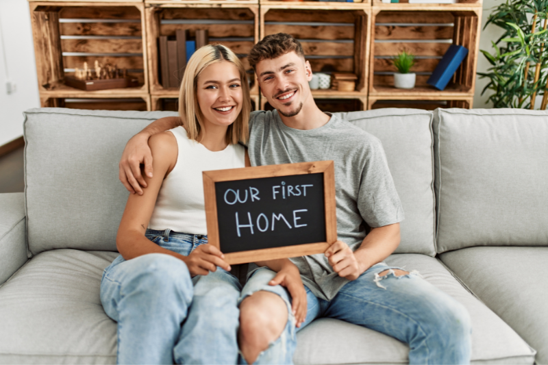 Preparing for a House Hunt is a couple of first-time homebuyers sitting on a couch, holding a small chalkboard sign that reads Our first home.